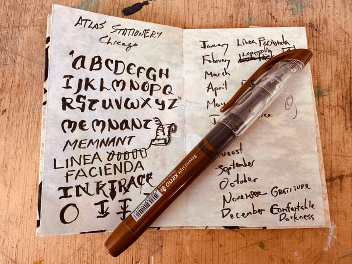 A small folded paper notebook with lettering and a monthly content schedule is open on a wood table with a brush pen resting on top.